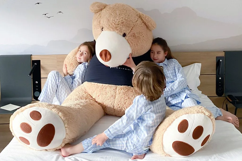 Deux enfant font un calin à un gros nounours en peluche dans un lit