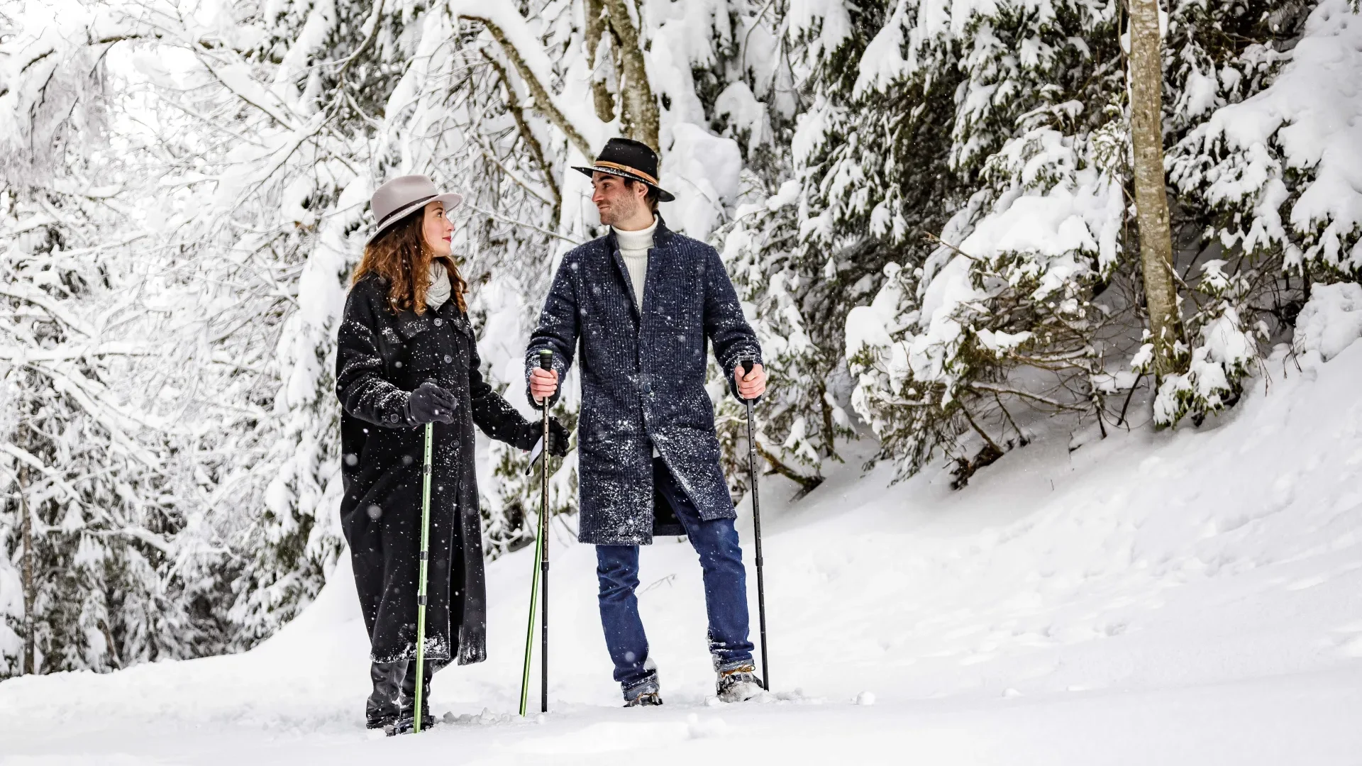 Elegante pareja con sombreros caminando en un paisaje nevado bajo los abetos.