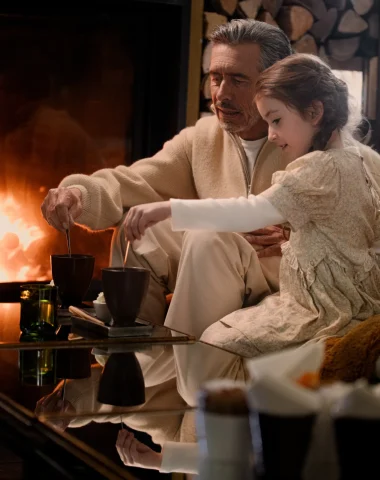 grand père et sa petite fille qui boivent un chocolat chaud devant un feu de cheminée