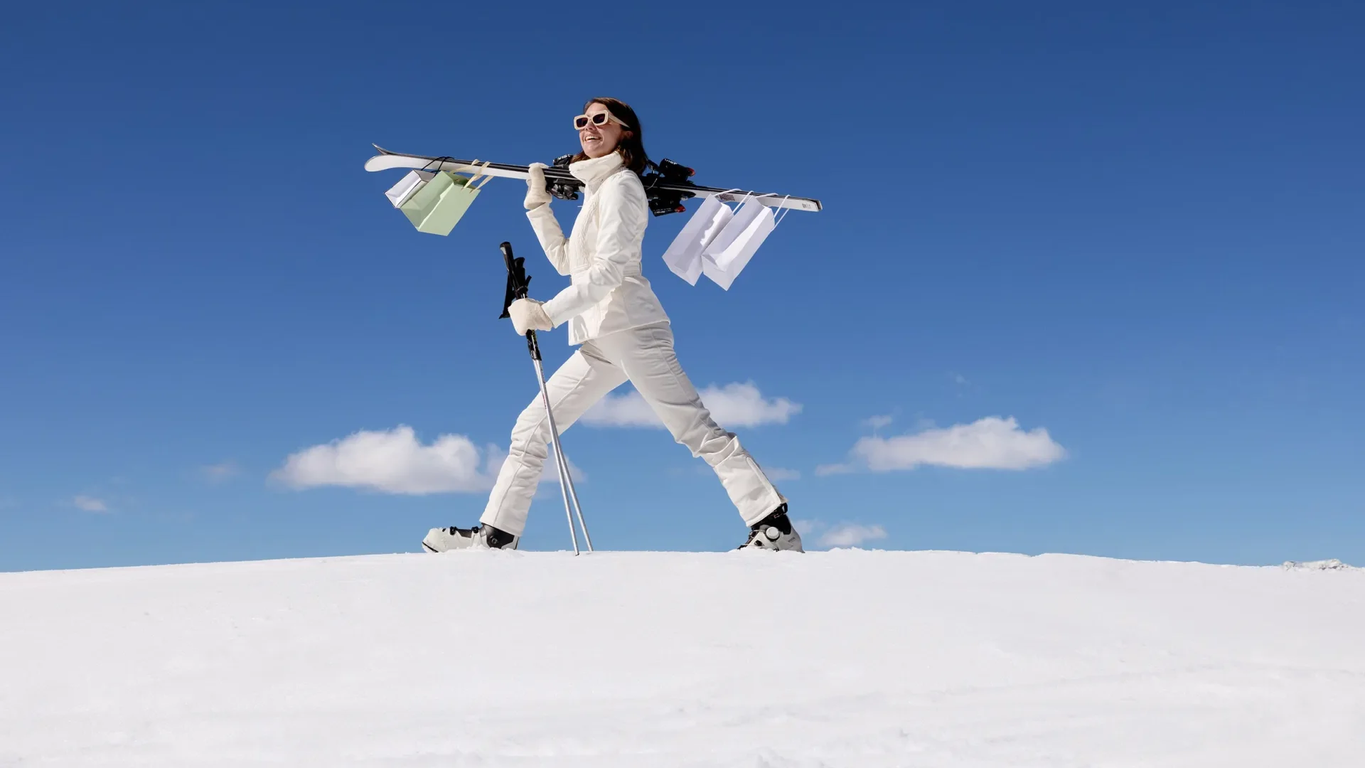 Jeune femme en tenue de ski qui marche sur les piste en tenant des batons, des skis et des sacs de shopping accrochés sur ses skis